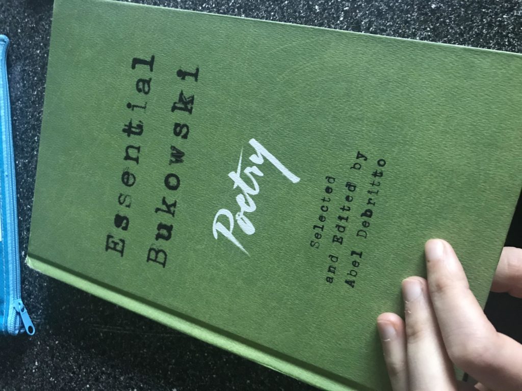 Green-covered book titled "Essential Bukowski Poetry," held by someone out of frame.