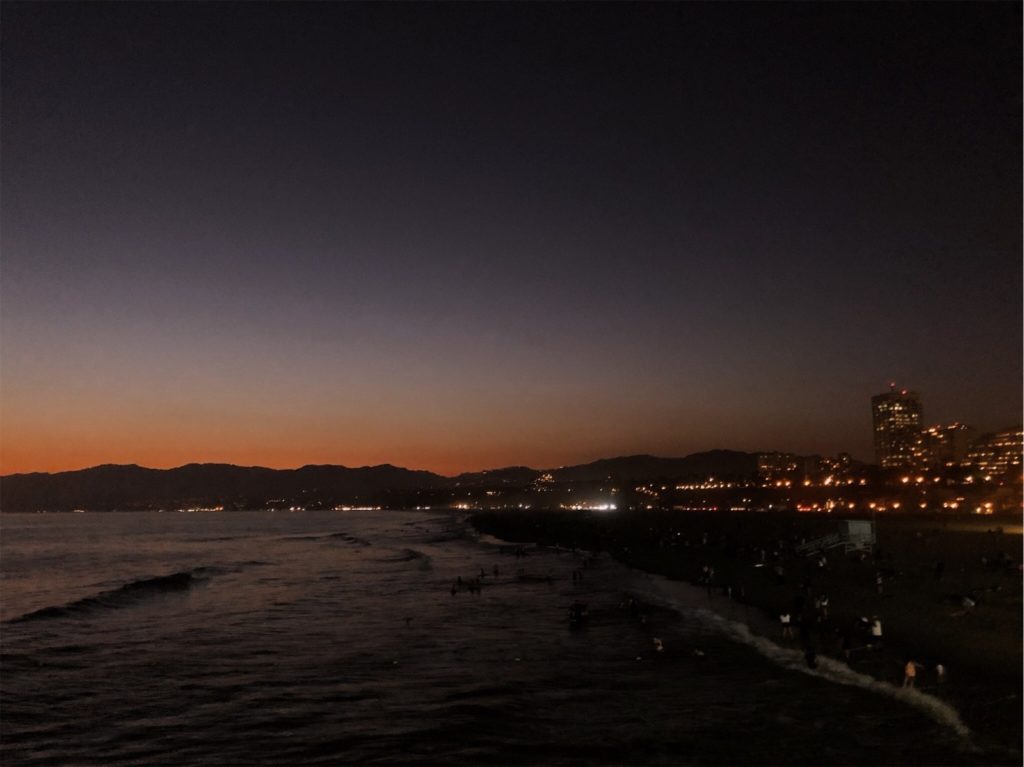 The sun is just below the horizon of the Santa Monica pier. The sky is orange near the horizon and deep blue at the top of the photo. The fading light reflects on the ocean in front of a city-lit pier.